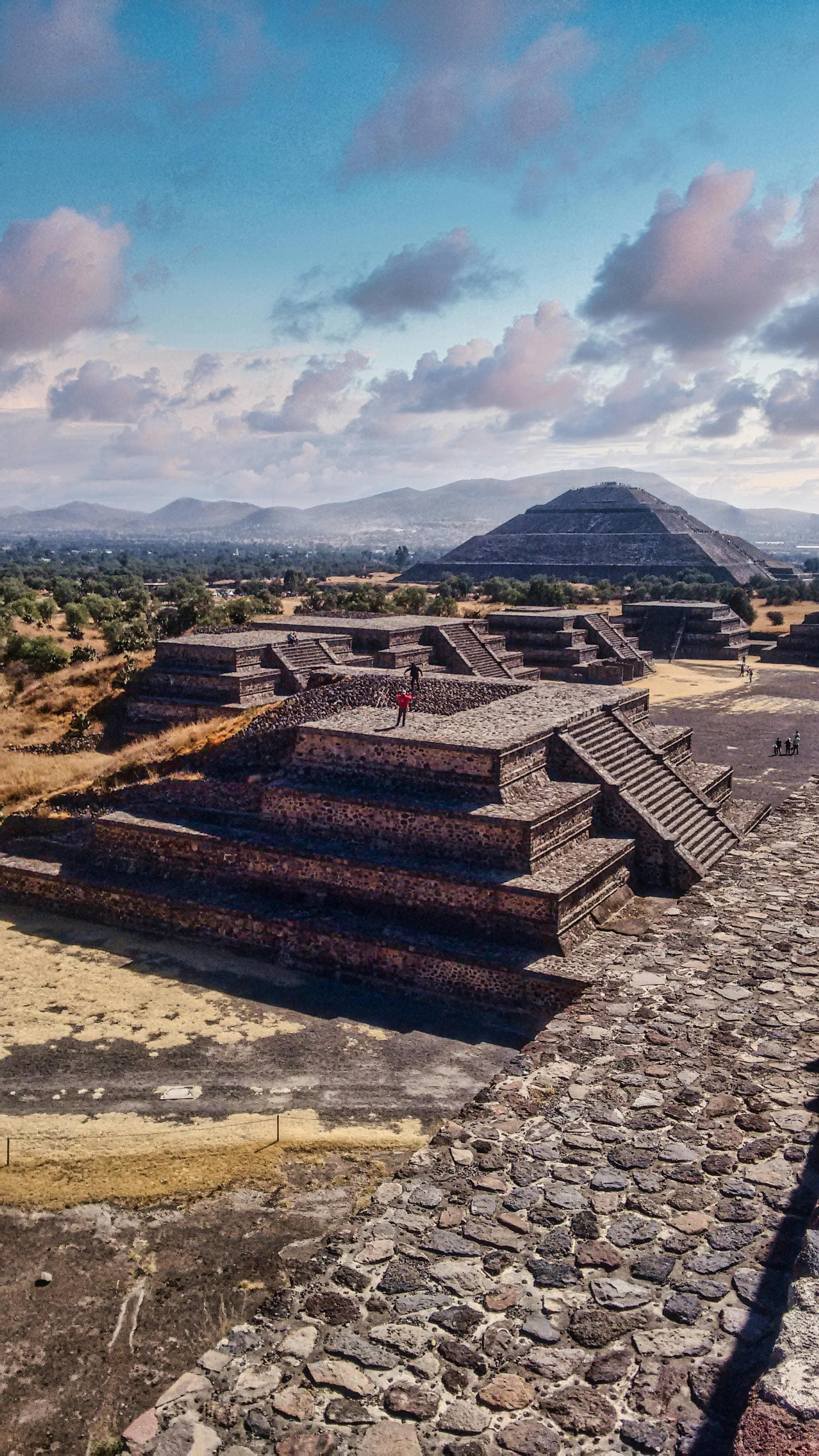 Ancient stone stepped pyramids at the Teotihuacan archaeological site under a cloudy blue sky.
